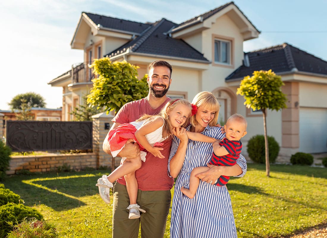 Personal Insurance - Portrait of a Young Mother and Father Holding Their Two Children as They Stand on the Green Grass in Front of Their Two Story Home